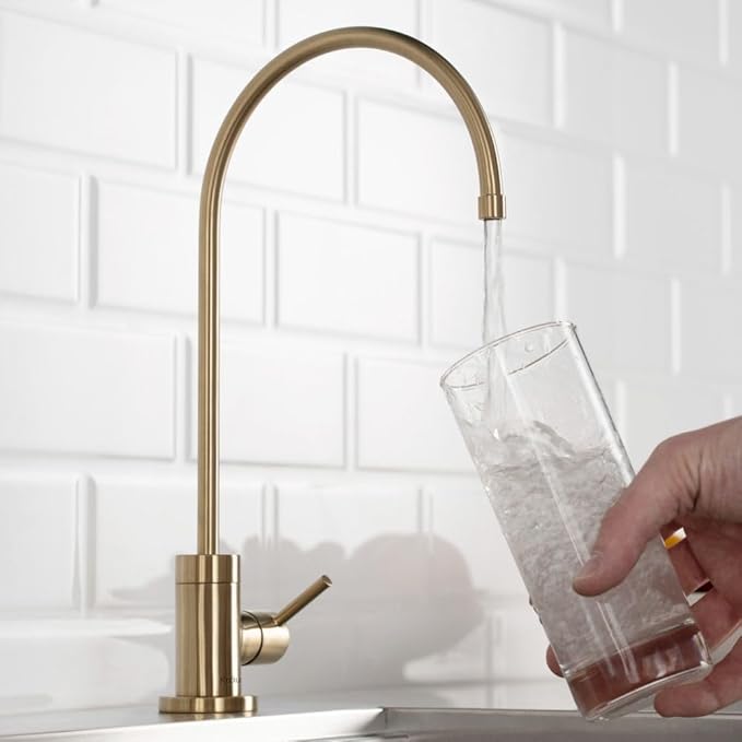 Gold kitchen faucet with a glass of water being filled against a white tiled wall.