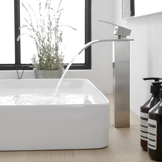 Modern bathroom sink with running water, plant, and bottles on a light wood floor.